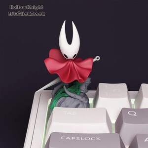Peut inclure: Figurine du personnage Hollow Knight, avec une tête blanche, une cape rouge et une épée, posée sur un clavier d'ordinateur. Le texte "Hollow Knight EduClickTrack" est visible. Objet de collection.