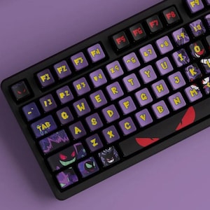 May include: A black mechanical keyboard featuring custom purple keycaps with yellow lettering. Several keys display graphic illustrations of a purple creature with red eyes. The spacebar features two red, almond-shaped eyes on a black background.