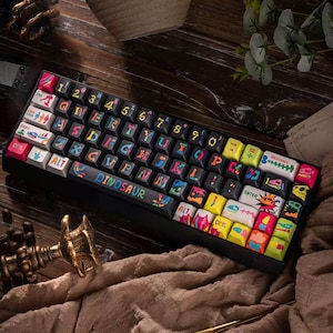 Puede incluir: Un teclado mecánico negro con teclas coloridas. Las teclas presentan varios diseños y la palabra "DINOSAUR" está impresa en la barra espaciadora. El teclado tiene un diseño compacto y está sobre una superficie de madera.