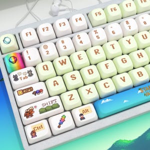 Può includere: Una tastiera personalizzata con tasti bianchi e verde chiaro. I tasti presentano vari disegni e testi in pixel art, tra cui "Tab", "Shift" e "Ctrl". La barra spaziatrice è blu con un design paesaggistico.