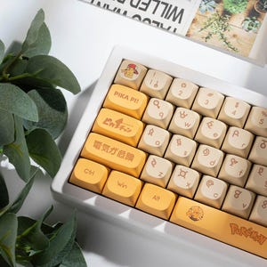 May include: A white mechanical keyboard with custom keycaps. The keycaps are a mix of cream and yellow, with Japanese text and Pikachu graphics. The keyboard has a compact layout and a white frame.
