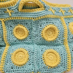 May include: Handmade crochet bag in a light blue and yellow color scheme. The bag features a repeating pattern of yellow circles on a light blue background, with yellow trim and handles. The crochet work is detailed and textured.