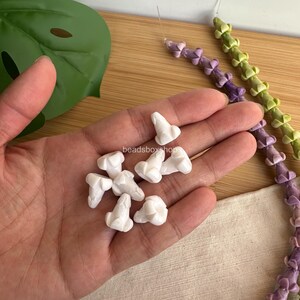 May include: A collection of small, white, flower-shaped beads with a central protrusion, held in a palm. Two long, flexible strands of similar beads in light purple and light green are visible nearby, featuring the text beadsboxshop in the center.