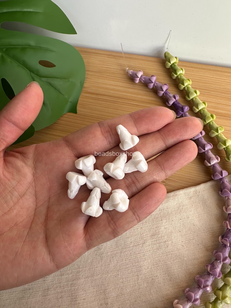 May include: A collection of small, white, flower-shaped beads with a central protrusion, held in a palm. Two long, flexible strands of similar beads in light purple and light green are visible nearby, featuring the text beadsboxshop in the center.