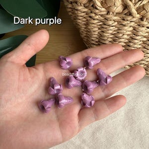 May include: A collection of small, dark purple beads shaped like flower buds with a smooth, glossy finish. Each bead features a central hole for threading. The text "Dark purple" and "beadsboxforhi" are visible in the image.