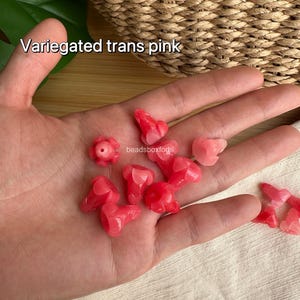 May include: A collection of small, variegated translucent pink beads shaped like flower buds. The beads feature a smooth, glossy finish and a central hole for stringing. The text "Variegated trans pink" and "beadsboxforhi" is visible in the image.