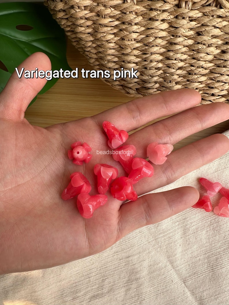 May include: A collection of small, variegated translucent pink beads shaped like flower buds. The beads feature a smooth, glossy finish and a central hole for stringing. The text "Variegated trans pink" and "beadsboxforhi" is visible in the image.