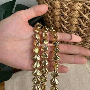 May include: A strand of metallic gold-toned beads, each shaped like a small seashell with detailed ridges. The beads are arranged in three vertical rows, reflecting light across their polished, curved surfaces. The text beadsboxforhi is visible.