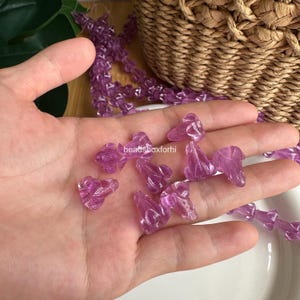 May include: A collection of translucent purple glass beads shaped like small flower buds. The beads are scattered across a palm and also appear as a continuous strand. The text beadsboxforhi is visible in the center of the image.