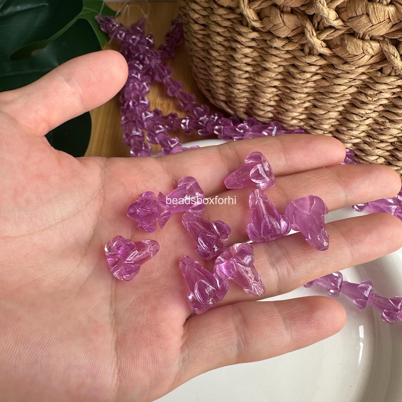 May include: A collection of translucent purple glass beads shaped like small flower buds. The beads are scattered across a palm and also appear as a continuous strand. The text beadsboxforhi is visible in the center of the image.