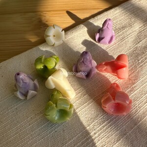 May include: A collection of small, flower-shaped beads in various pastel colors including lavender, cream, light green, and coral. Each bead features a central hole and sculpted petal details. The text beadsboxshop is visible in the center.