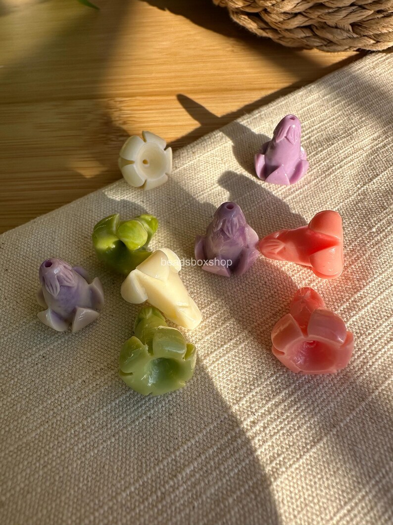 May include: A collection of small, flower-shaped beads in various pastel colors including lavender, cream, light green, and coral. Each bead features a central hole and sculpted petal details. The text beadsboxshop is visible in the center.