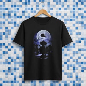 Op de afbeelding: Zwart T-shirt met een Halloween-thema. Het ontwerp toont een volle maan, een heks op een bezem en silhouetten van bomen en een decoratieve structuur. Het T-shirt hangt aan een houten hanger.