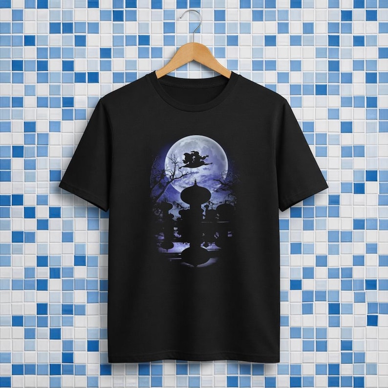 Op de afbeelding: Zwart T-shirt met een Halloween-thema. Het ontwerp toont een volle maan, een heks op een bezem en silhouetten van bomen en een decoratieve structuur. Het T-shirt hangt aan een houten hanger.