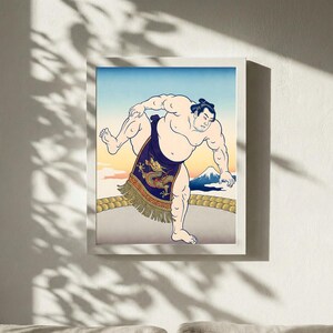 May include: Framed print depicting a sumo wrestler. The artwork shows a wrestler in a traditional mawashi with a dragon design. The background features a mountain and a gradient sky, within a white frame.