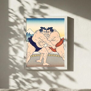 May include: Framed print depicting a traditional Japanese sumo wrestling scene. Two wrestlers grapple, wearing blue and red loincloths. The background shows a mountain and a gradient sky, within a white frame.