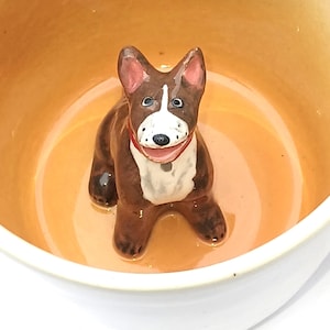 Könnte beinhalten: Eine Keramik-Hundefigur mit braunen und weißen Markierungen und einem roten Halsband sitzt in einer hellorangen Schüssel. Der Hund hat einen fröhlichen Gesichtsausdruck. Die Schüssel hat eine weiße Außenseite.