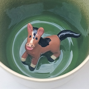 Può includere: Una tazza in ceramica con interno verde ed esterno bianco. All'interno della tazza c'è una statuina di cavallo marrone e bianco con criniera e coda nere. Il cavallo è in posa giocosa, aggiungendo un tocco stravagante alla tazza.
