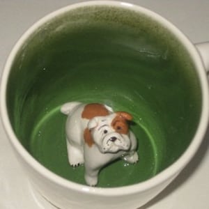 Könnte beinhalten: Weißer Keramikbecher mit grünem Innenraum, mit einer 3D-Bulldoggenfigur. Die Bulldogge ist weiß mit braunen Flecken und scheint in der Tasse zu sitzen. Der Becher hat einen weißen Henkel und eine glänzende Oberfläche.