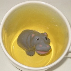 May include: A white ceramic mug with a yellow interior. A gray hippo figurine is centered inside the mug. The hippo has pink accents on its mouth and ears. The mug is viewed from a top-down perspective.