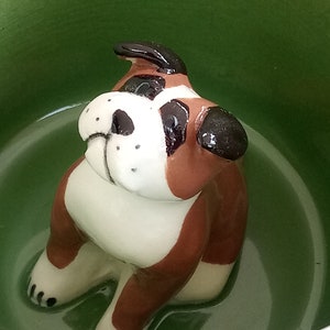 Könnte beinhalten: Eine Keramik-Bulldoggen-Figur mit braunen, weißen und schwarzen Markierungen sitzt in einer grünen Schüssel. Die Bulldogge hat ein detailliertes Gesicht und ist so positioniert, als würde sie sitzen. Die Schüssel hat eine glänzende Oberfläche.