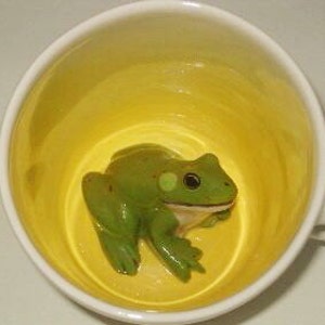 May include: A white ceramic mug with a bright yellow interior. A green frog figurine with a light green spot on its side rests inside the mug. The frog has black eyes and a white belly. The mug is empty.