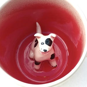 May include: A white ceramic mug with a red interior. A small, white ceramic dog with black spots is sitting inside the mug.