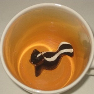 Könnte beinhalten: Eine weiße Keramiktasse mit orangefarbenem Innenraum. In der Tasse befindet sich eine kleine, dreidimensionale Stinktierfigur mit schwarz-weißen Streifen. Das Stinktier blickt nach links.