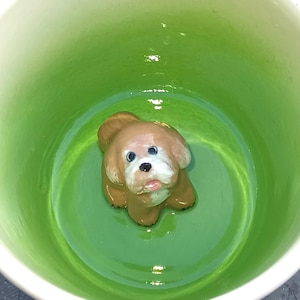 Op de afbeelding: Een keramieken hondenfiguur in een groene beker. De hond is bruin met witte accenten en blauwe ogen. De beker heeft een glanzende afwerking.