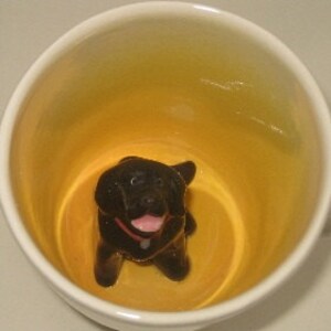 Könnte beinhalten: Eine weiße Keramiktasse mit einer schwarzen Labrador Retriever-Figur im Inneren. Der Hund hat ein rosa Halsband und Zunge, vor einem gelben Innenraum. Die Tasse wird von oben betrachtet.