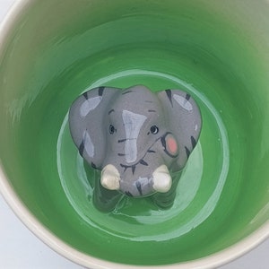 Könnte beinhalten: Eine Keramik-Elefantenfigur in Grau und Weiß, die in einer grün-weißen Keramikschale sitzt.