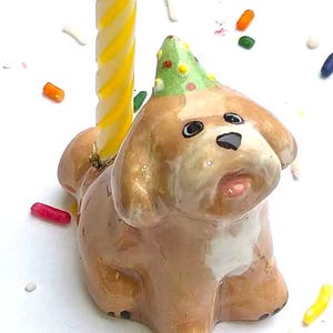 May include: A ceramic dog-shaped candle holder with a yellow and white striped candle. The dog is brown and white, wearing a green party hat with colorful dots. Colorful sprinkles are scattered around the base.