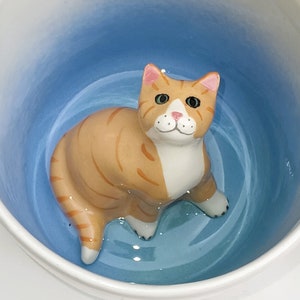 Könnte beinhalten: Eine orangefarbene Tabby-Katzenfigur aus Keramik in einer blau-weißen Schüssel. Die Katze hat grüne Augen, eine rosa Nase und weiße Pfoten. Die Schüssel hat einen blau verlaufenden Innenraum und einen weißen Rand.