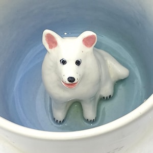 Könnte beinhalten: Weißer Keramikbecher mit einem 3D-modellierten Samojeden-Hund im Inneren. Der Hund hat rosa Ohren, schwarze Augen und eine schwarze Nase. Das Innere des Bechers hat einen blau-weißen Farbverlauf.