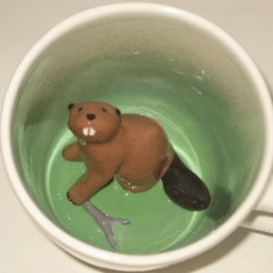 Puede incluir: Una taza de cerámica blanca con interior verde. Dentro, una figura de castor marrón con dientes blancos y cola negra sostiene una ramita gris. La taza está sobre una superficie blanca.