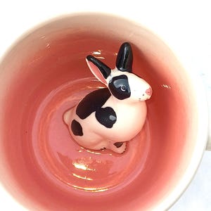 Könnte beinhalten: Eine weiße Keramiktasse mit rosafarbenem Innenraum. Eine schwarz-weiße Hasenfigur schaut aus der Tasse.