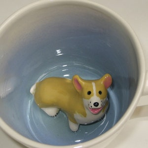 Könnte beinhalten: Weiße Keramiktasse mit hellblauem Innenraum und einem geformten Corgi-Hund im Inneren. Der Corgi ist gelb, weiß und rosa, mit einem lächelnden Gesichtsausdruck. Die Tasse hat einen weißen Griff.