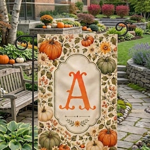 May include: A rectangular garden flag featuring a central orange letter A inside an ornate frame. The design includes illustrations of orange, green, and white pumpkins, autumn leaves, berries, and yellow flowers on a cream-colored background.