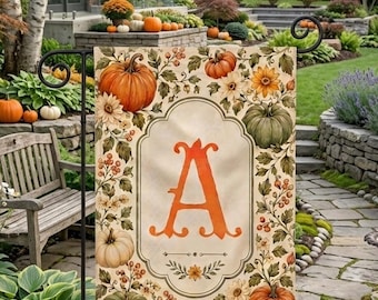 Könnte beinhalten: Eine rechteckige Gartenflagge mit einem zentralen orangefarbenen Buchstaben A in einem verzierten Rahmen. Das Design zeigt Illustrationen von orangefarbenen, grünen und weißen Kürbissen, Herbstblättern, Beeren und gelben Blumen auf cremefarbenem Grund.