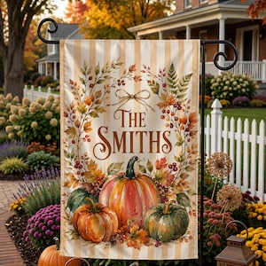 May include: A rectangular garden flag featuring a vertical beige and white striped background. The design includes a central wreath of autumn leaves and berries surrounding the text "THE SMITHS" above three pumpkins in orange and green hues.