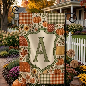 May include: A rectangular garden flag featuring a patchwork design of orange and green plaid, polka dots, and autumn leaves. A central cream-colored shield with a decorative green border displays a large, ornate green letter A in the center.