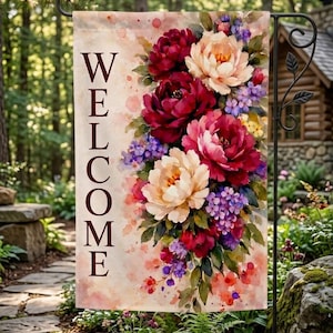 May include: A garden flag with the word "WELCOME" in brown capital letters. The flag features a watercolor floral design with burgundy, pink, and purple flowers. The flag is hanging on a black metal stand in a garden setting.