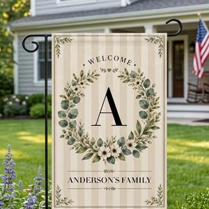 May include: A rectangular garden flag featuring a beige and cream vertical striped background. The design includes a central floral wreath with green leaves and white flowers surrounding the letter A. Text reads WELCOME and ANDERSON'S FAMILY.
