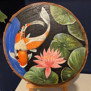 May include: A hand-painted artwork on a wooden slice, depicting a koi carp and water lily. The image shows an orange and white koi fish in blue water, a pink water lily, and green lily pads against a black backdrop. The wood slice is circular.