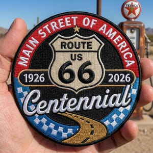 May include: A circular embroidered patch featuring a black background with a central Route 66 shield. The text reads Main Street of America, 1926, 2026, and Centennial. The design includes a gold road, blue checkered border, and red outer ring.