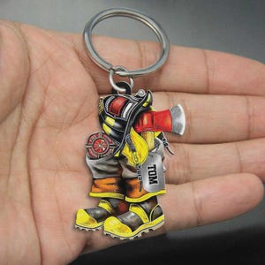 May include: A firefighter-themed keychain featuring a detailed design of a firefighter's boots, helmet, and axe. The keychain includes a silver keyring and a tag that reads "TOM". The boots are yellow and black.