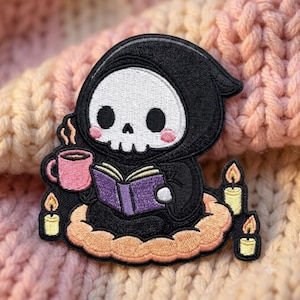 May include: An embroidered patch featuring a black-hooded skeleton character with a white skull and pink cheeks. The character holds a pink mug with steam and a purple book while sitting on a peach-colored cloud surrounded by three small yellow candles.