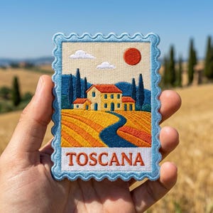 May include: A rectangular embroidered patch with a scalloped edge featuring a landscape of a yellow house, cypress trees, and rolling fields under a red sun. The sky is cream-colored with two white clouds. The word TOSCANA is stitched in red.