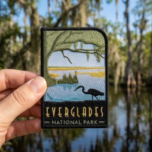 May include: A rectangular embroidered patch featuring a landscape of the Everglades National Park. The design shows a light blue sky, a yellow marsh, and a dark silhouette of a bird. The text EVERGLADES NATIONAL PARK is stitched in gold.