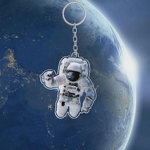May include: A keychain featuring an astronaut in a white spacesuit, with the name "JONKSON" printed on the suit. The astronaut is suspended from a silver chain and ring, set against a backdrop of Earth and space.
